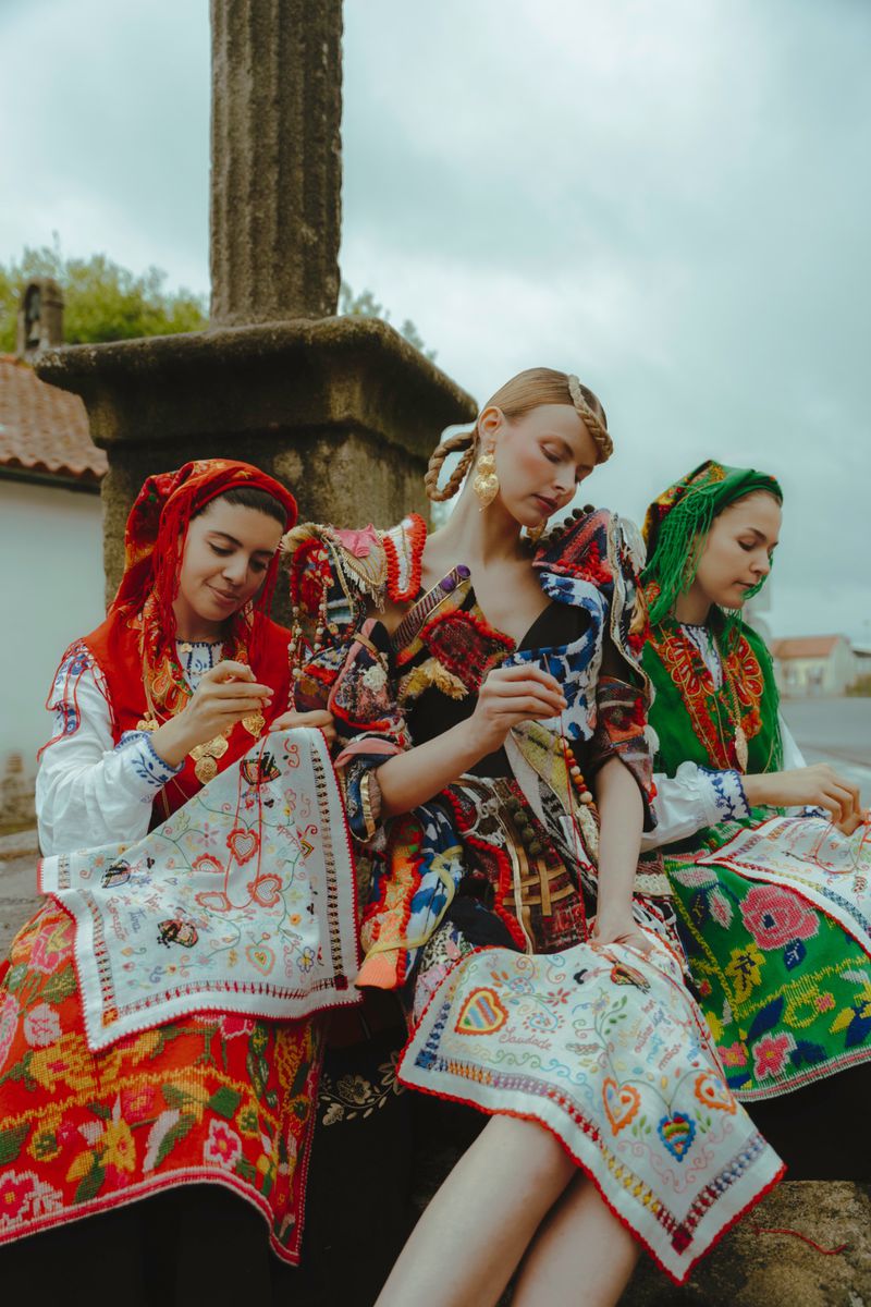 Three adult women in traditional clothing sit near a stone pillar in an outdoor setting. Embroidery work on cloths is visible.