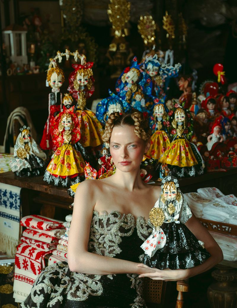 Woman in ornate dress holding a vintage doll amid a collection of antique dolls, creating a nostalgic and artistic composition.