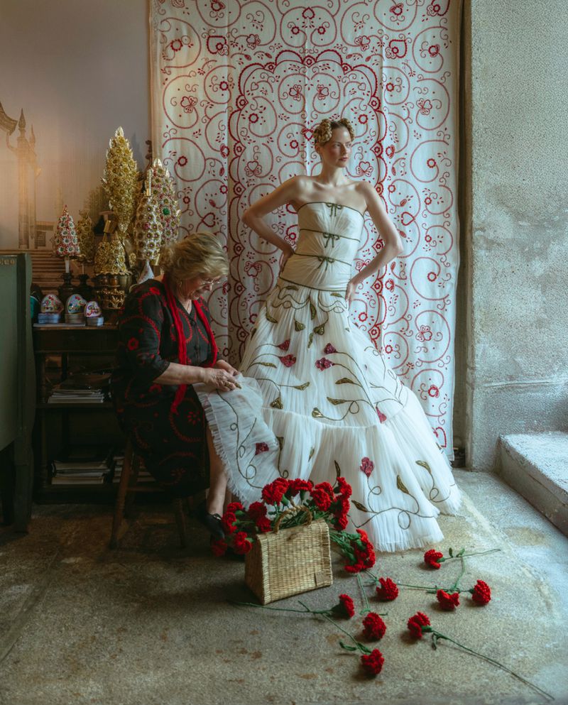 Woman in strapless white gown poses beside seamstress and red roses with ornate background. Fashion editorial with vintage aesthetic.