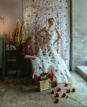 Woman in strapless white gown poses beside seamstress and red roses with ornate background. Fashion editorial with vintage aesthetic.
