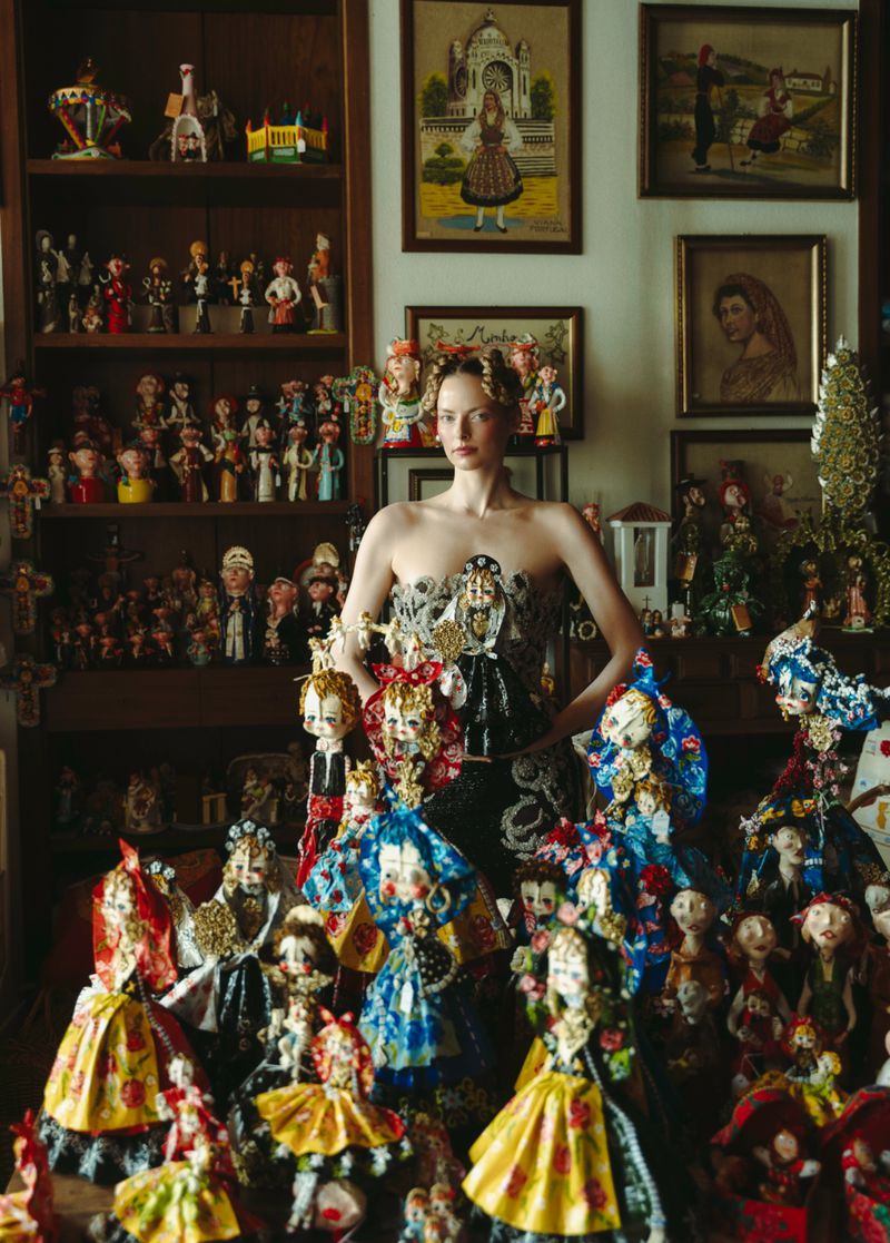 A fashion portrait presents a woman adorned in a strapless bodice-style dress surrounded by an array of handcrafted dolls in an indoor setting.
