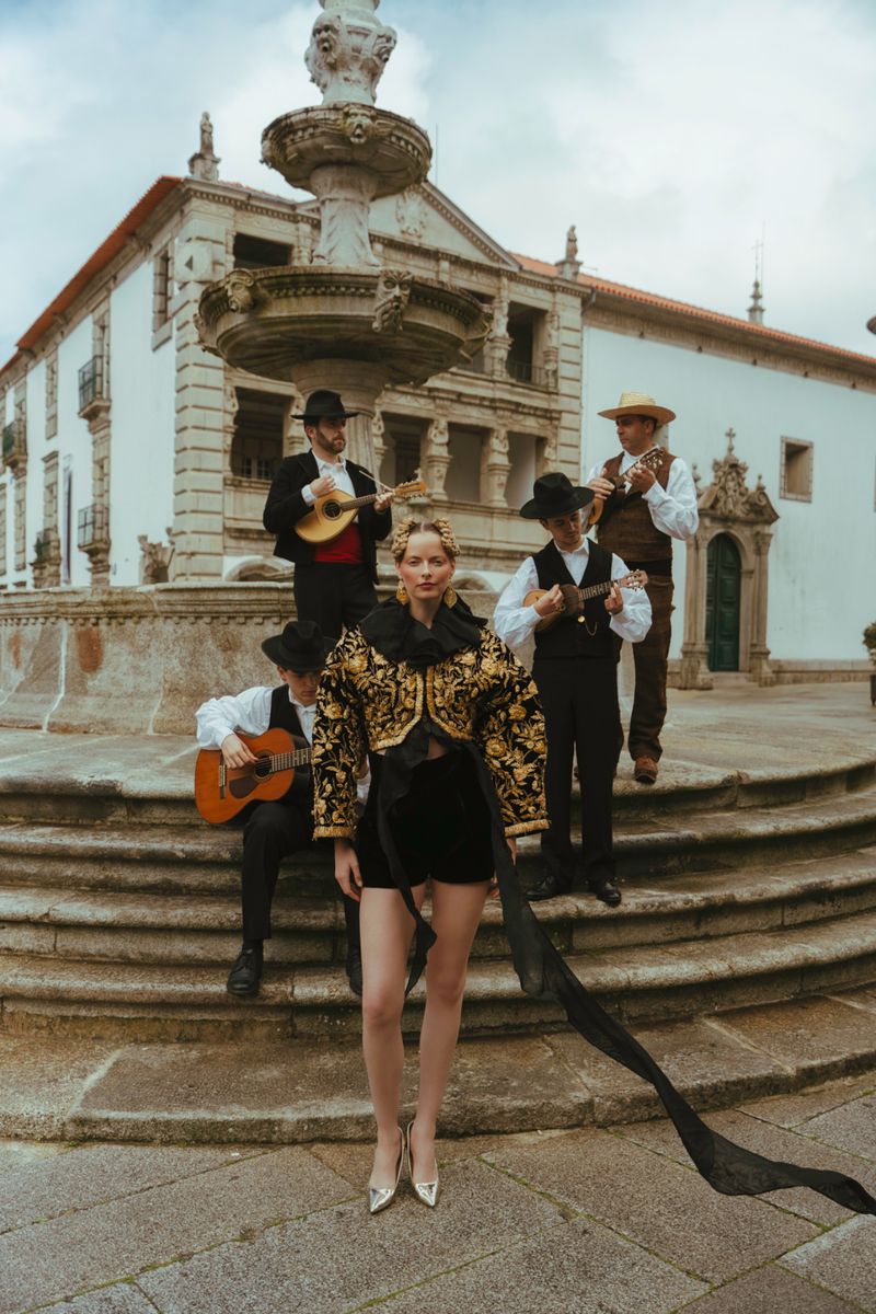 Fashionable woman poses on steps near a fountain, wearing an ornate jacket, with musicians in the background in an outdoor setting.