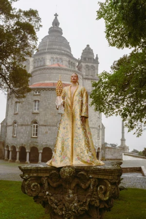 Woman in elaborate gold coat stands on stone plinth with church backdrop. Exterior shot in muted light shows architecture, fashion.