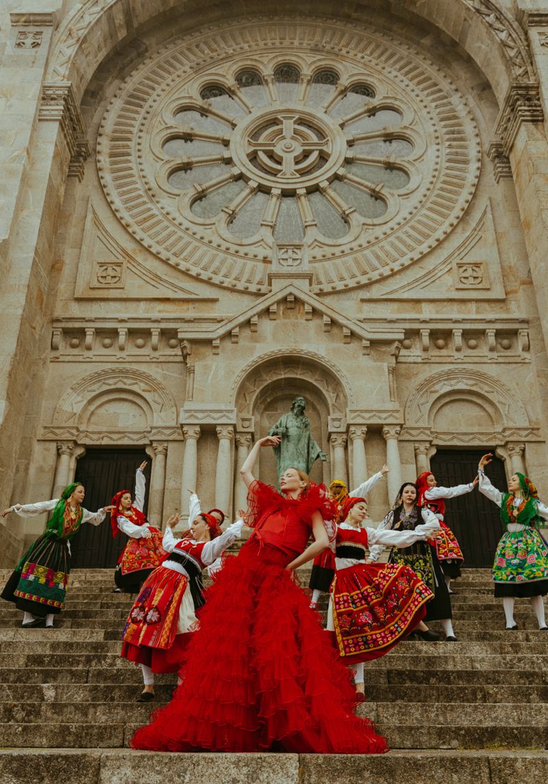 Group of women in traditional dresses dancing on the steps of a stone church. A woman in a long red dress stands out in the foreground.