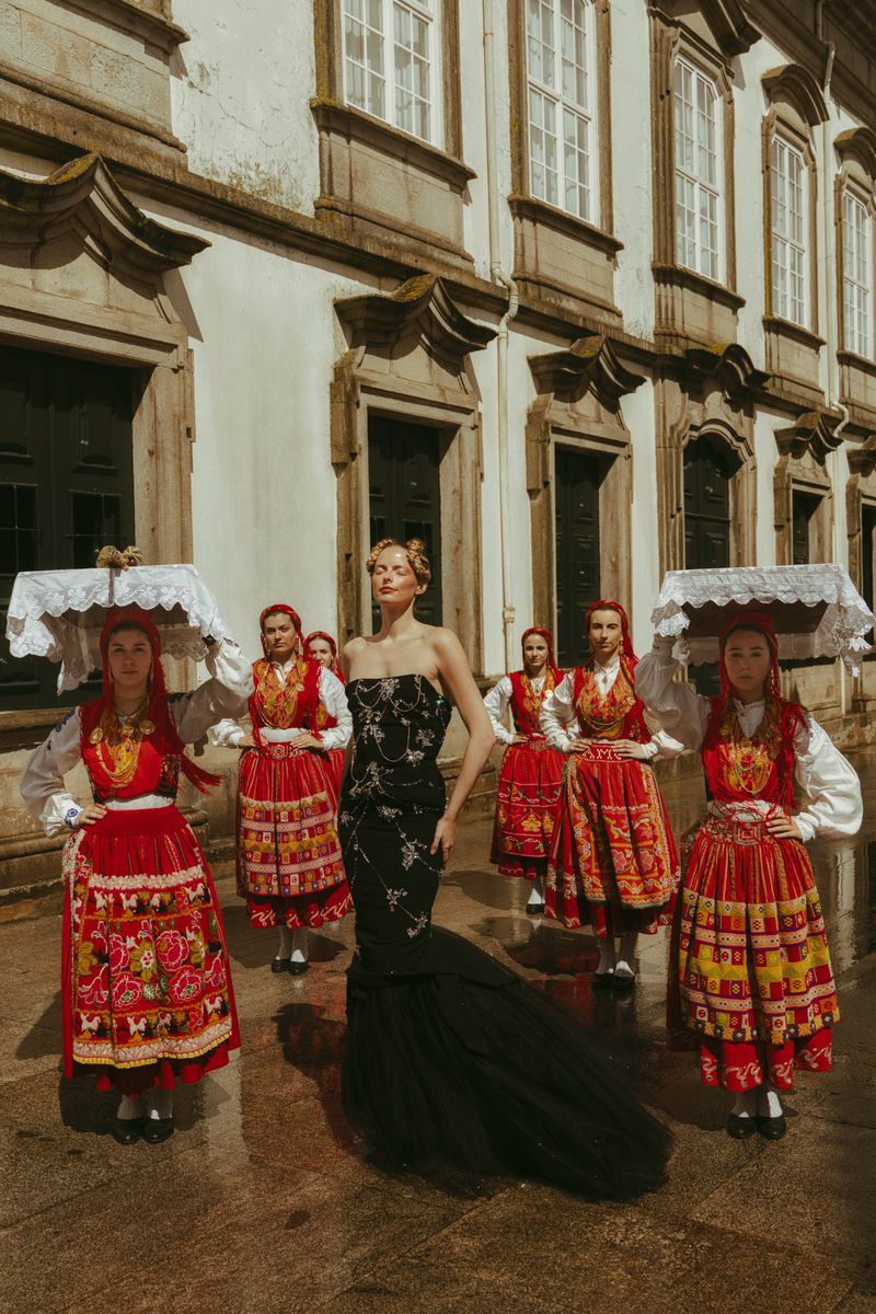 Fashion editorial with model in black gown surrounded by women in traditional red folk dresses, set against a stone building backdrop.