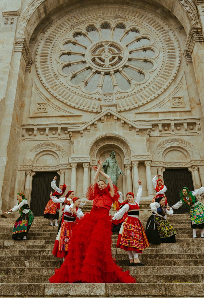 Woman in red gown surrounded by a group of women in folk costumes on steps of stone building with a rose window and statue.