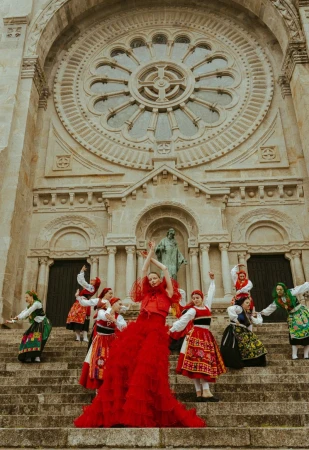 Woman in red gown surrounded by a group of women in folk costumes on steps of stone building with a rose window and statue.