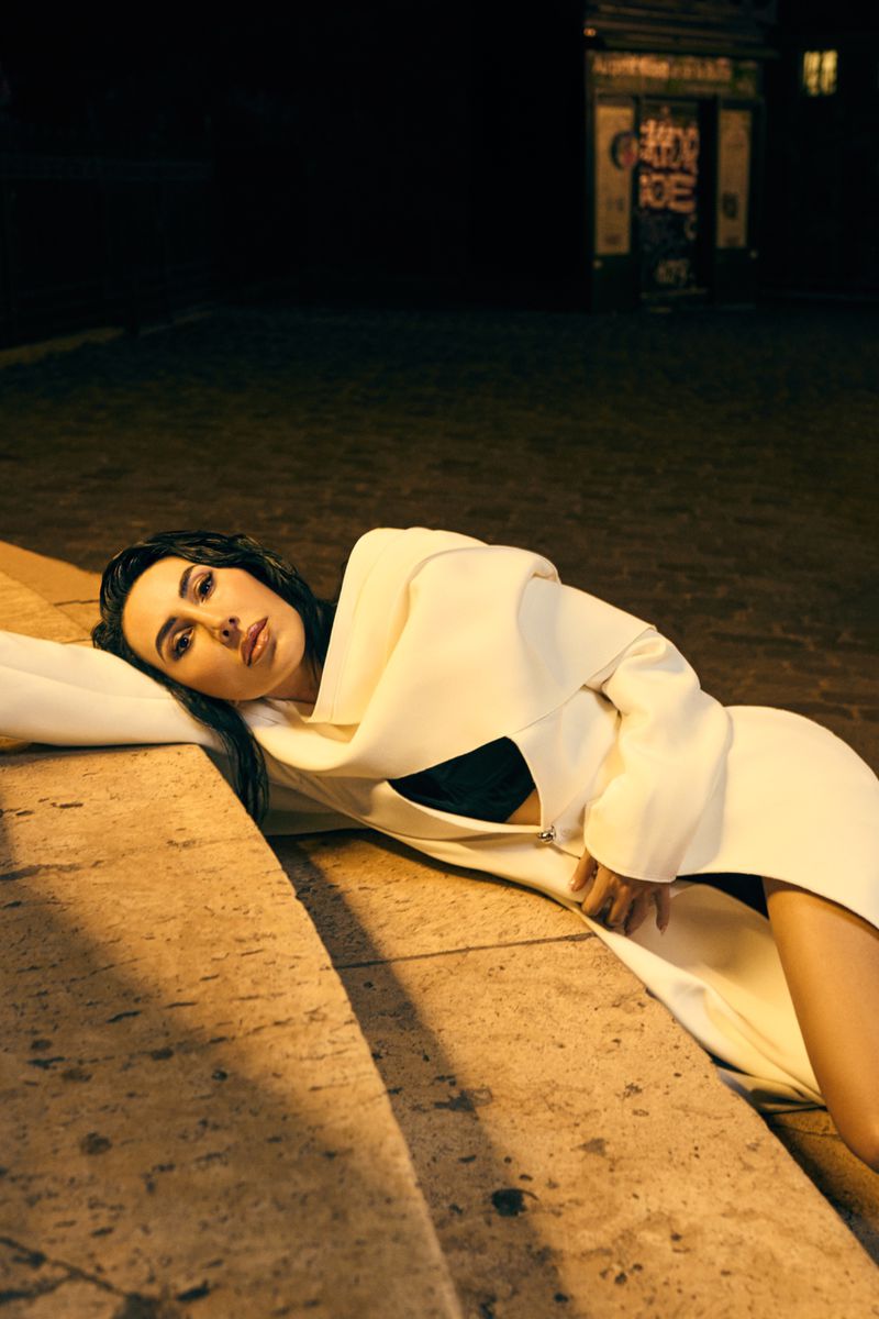 Adult woman in an off-white coat poses in a reclined position outside at night with shadowed stairs in the foreground.