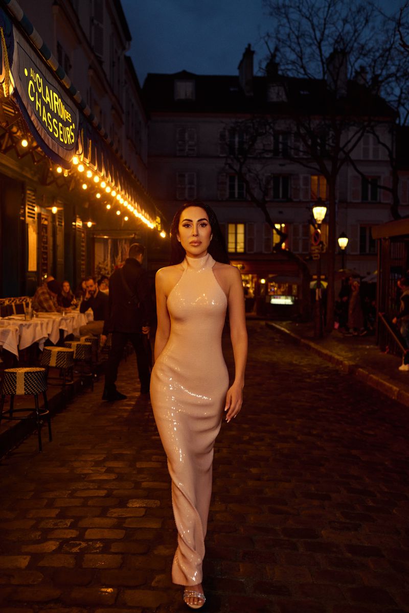 A brunette woman in a sequined gown walks a cobblestone street in front of a restaurant with warm lighting in an urban setting at dusk.