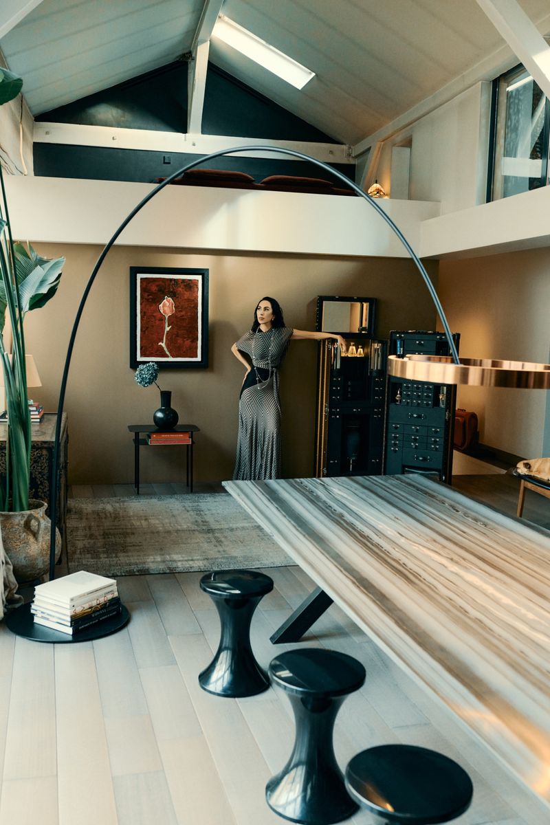 A woman in an elegant dress stands inside a modern home featuring a marble table, black stools, and contemporary decor.