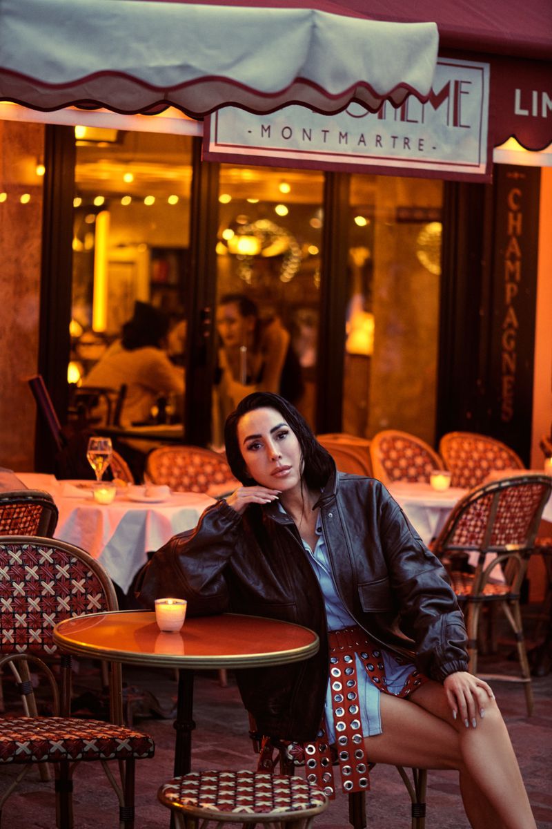 Woman in a leather jacket sits at a table outside a cafe with a glass of champagne, with warm lighting from the restaurant interior.