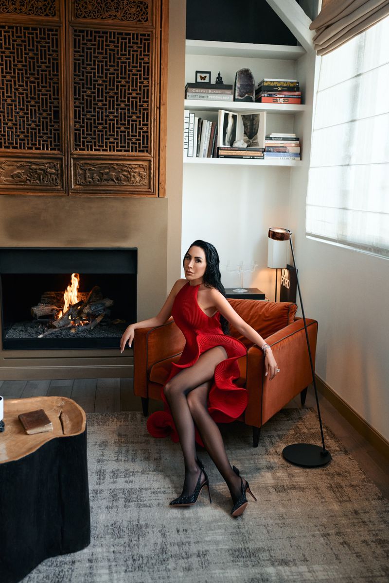 A brunette woman in a red dress and black hosiery sits in an orange armchair in a stylish room with a fireplace and modern decor.
