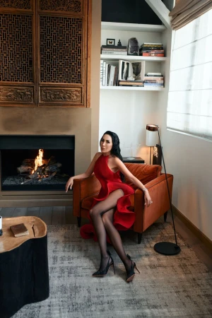 A brunette woman in a red dress and black hosiery sits in an orange armchair in a stylish room with a fireplace and modern decor.