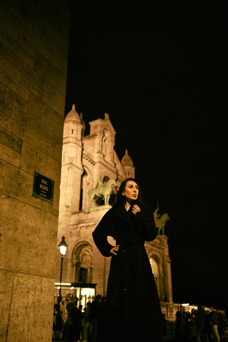 Adult woman wearing a dark coat stands in front of the Sacré-Cœur basilica in Paris at night, creating an elegant fashion portrait.