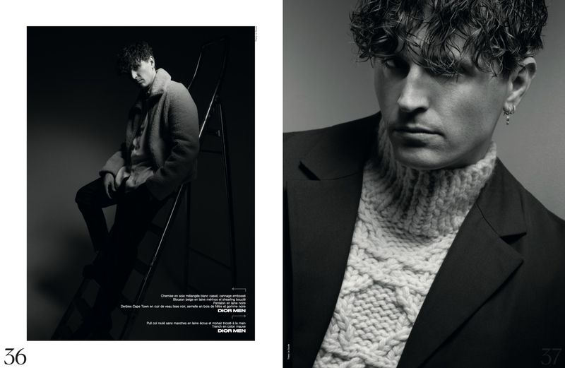 Black-and-white fashion shot of a curly-haired adult male model wearing a turtleneck sweater and blazer, sitting on a ladder in a studio.