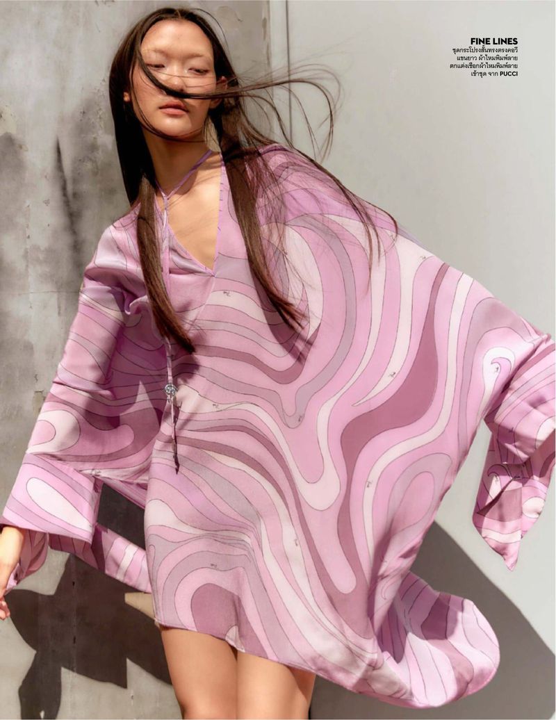A woman poses in a lilac dress with wavy patterns. Wind blows her dark hair. Natural lighting. The background is a plain, light-colored wall.