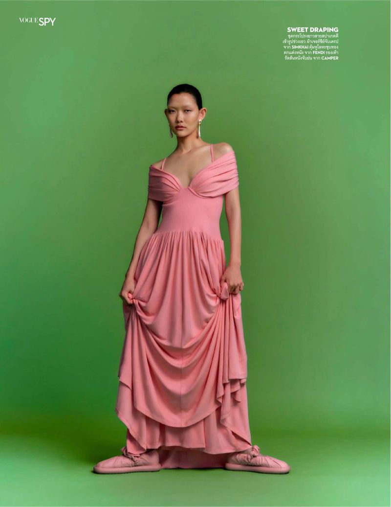 Woman in a pink gown posing against a green background. The dress is draped and flowing. She wears earrings and matching shoes.