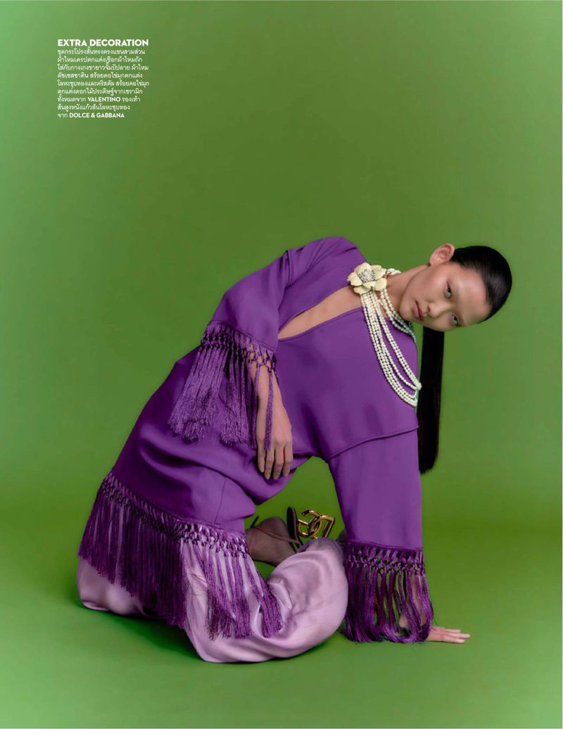 Fashion shot of an Asian woman in purple fringe dress with pearl necklace, kneeling in front of a solid green backdrop.