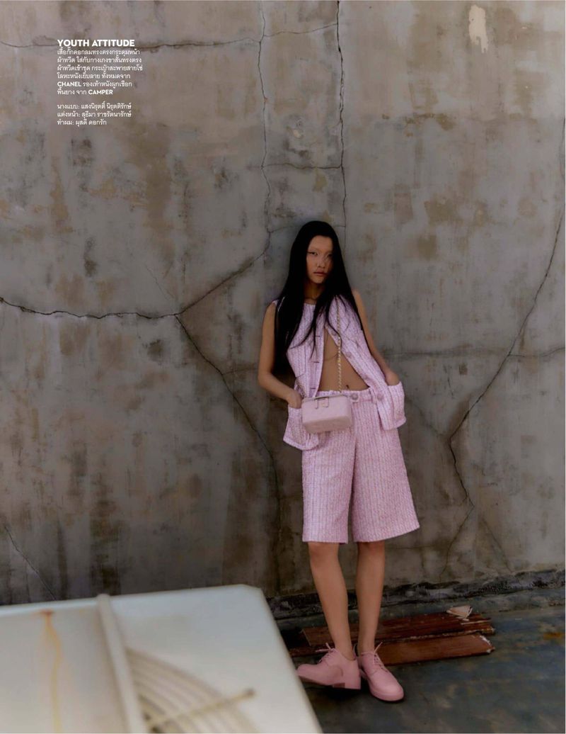 Fashion editorial shot of a young Asian woman in a pink shorts suit posing against a textured grey wall with accessories and pink shoes.