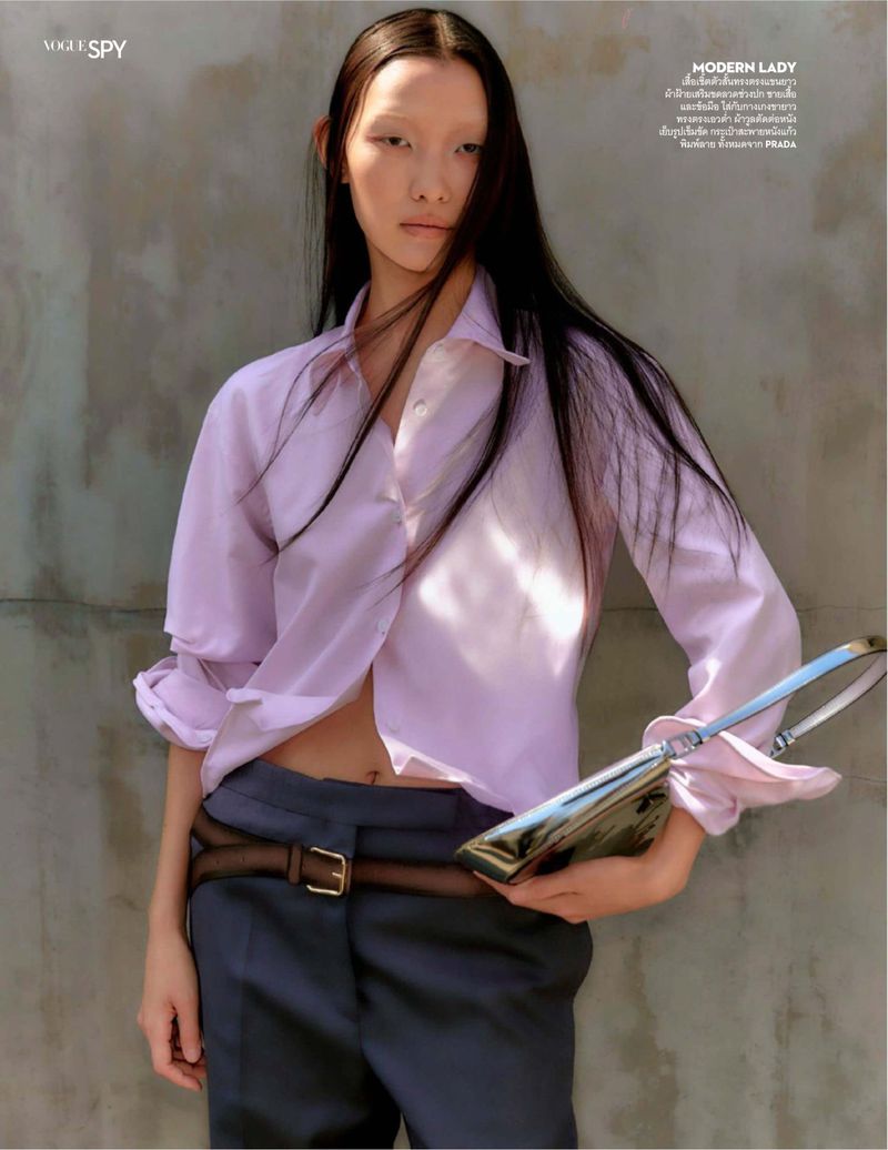 Fashion editorial shot: Asian woman with long hair poses in lilac blouse, grey pants, and brown belt, holding a metallic purse.