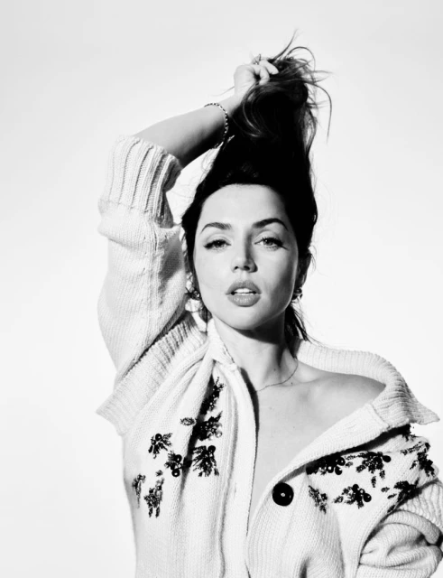 Black and white portrait of Ana de Armas holding her hair, wearing earrings and a patterned cardigan against a white background.