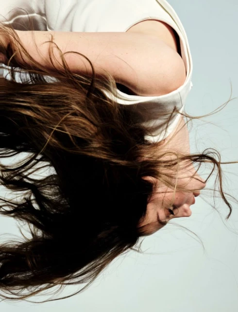 Woman with long brown hair hangs upside down wearing a white shirt against a pale blue background in a portrait orientation.