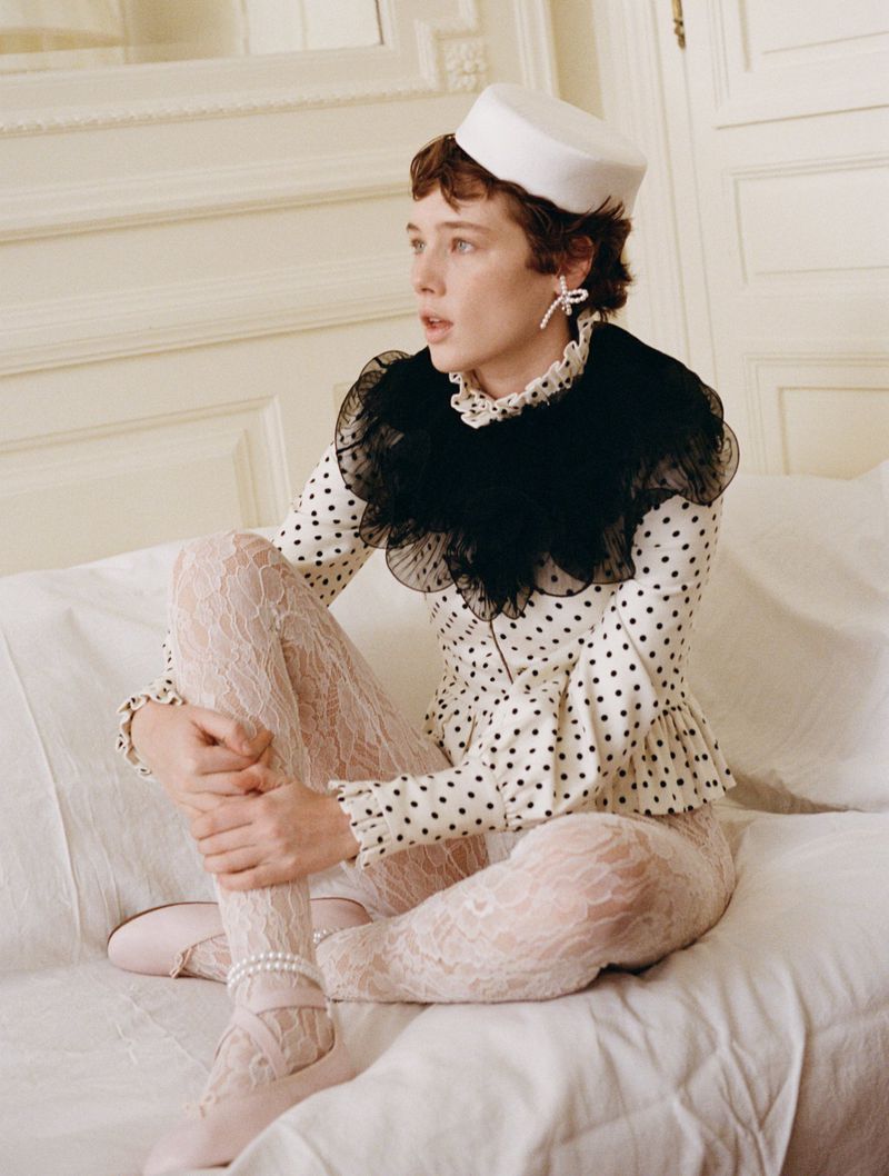 Young woman in vintage-inspired polka dot top, white tights, and hat poses seated in a light, vintage-decorated interior.