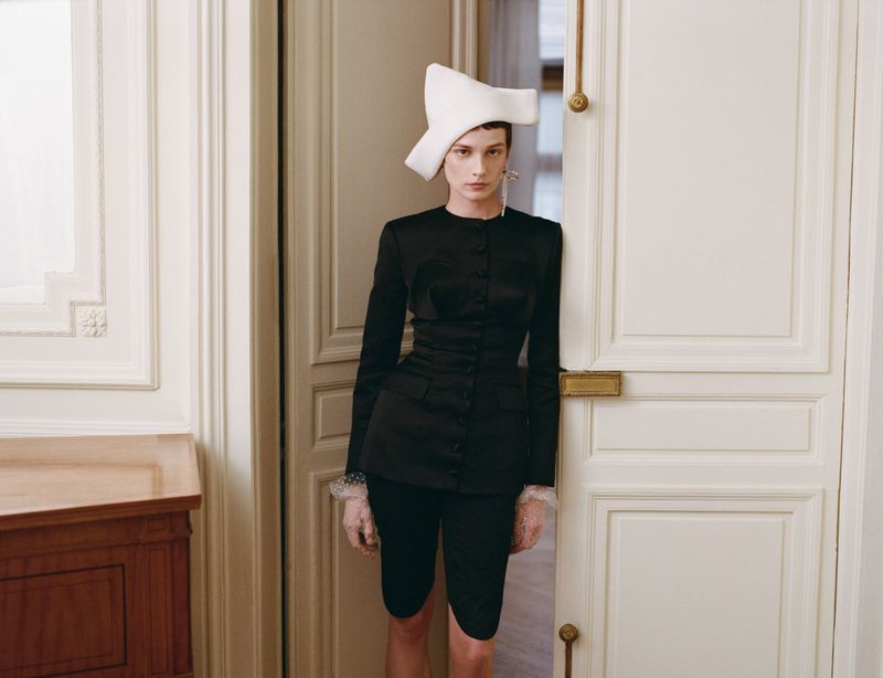 Elegant woman in black jacket, matching capri pants, and a structured white hat posing in a bright room next to a white door.