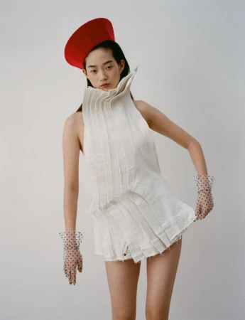 Fashion portrait of Asian woman in white pleated outfit and red hat against white backdrop, wearing sheer polka dot gloves.