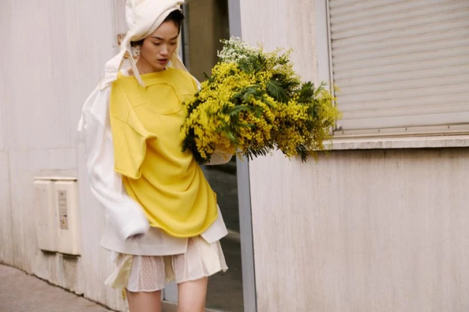 A woman walks outdoors holding a bouquet of yellow flowers, wearing an oversized yellow shirt and white skirt, with white head covering.