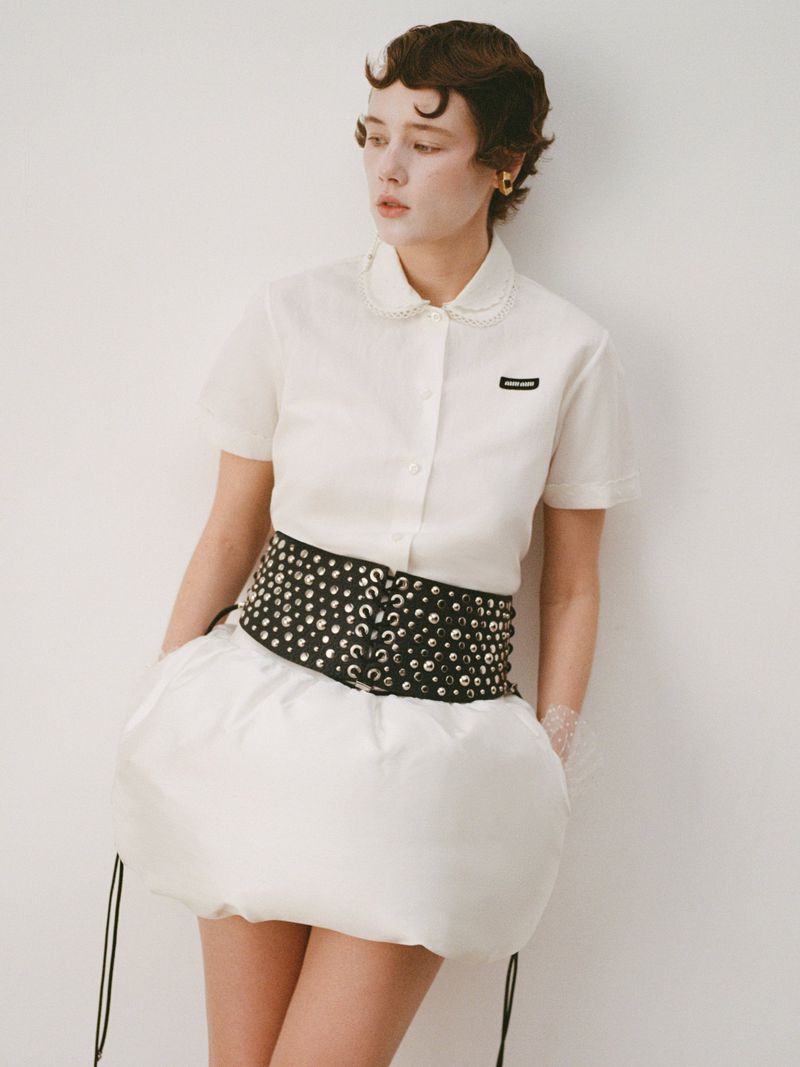 A woman with short, styled hair in a collared shirt and distinctive skirt poses in a studio setting with a neutral background.