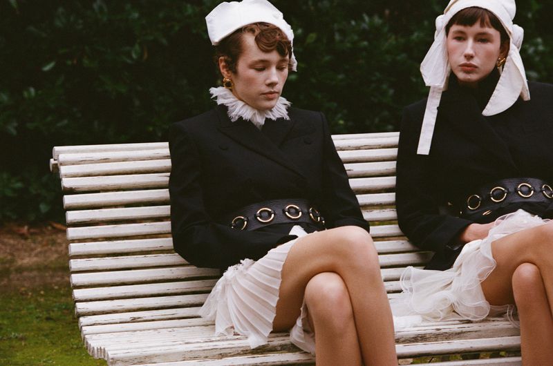 Two women in black blazers, white skirts, and white headwear sit on a white bench in a lush outdoor setting, captured in a medium shot.