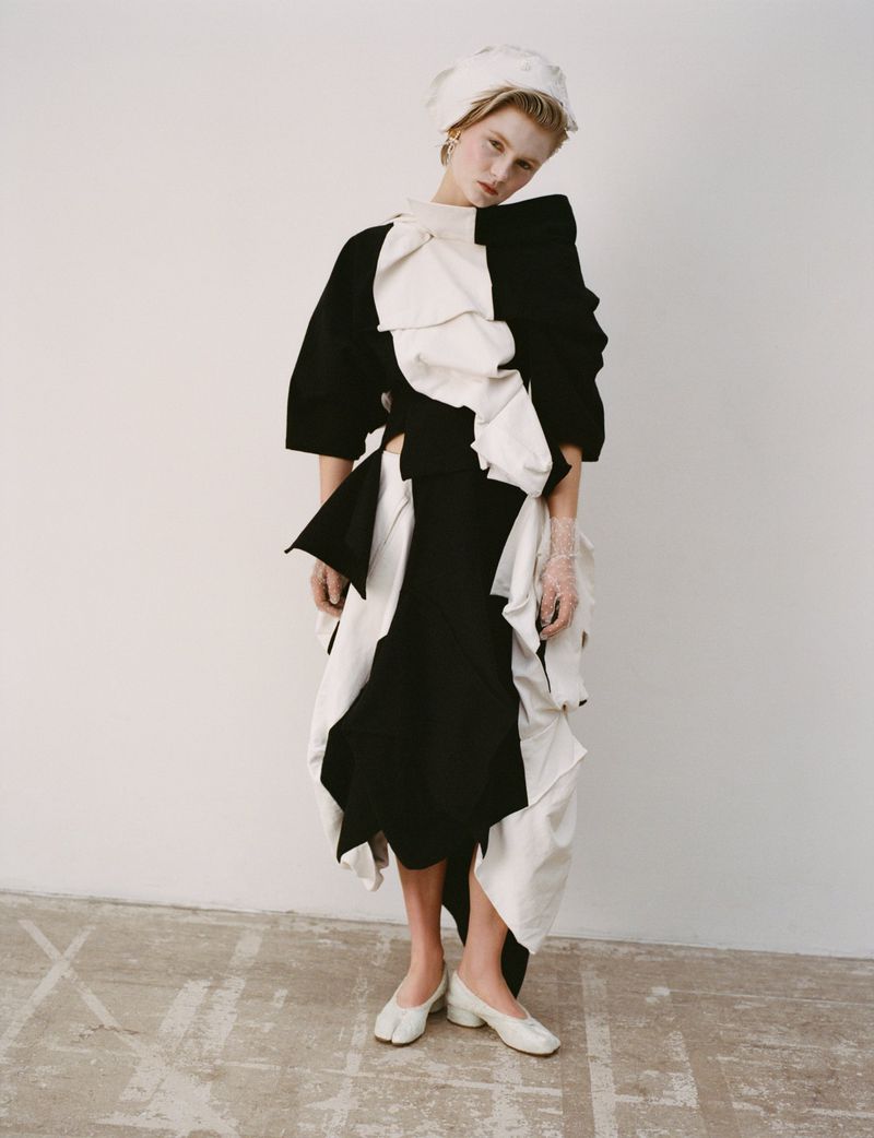 A young woman with blonde hair poses in a studio wearing a black-and-white dress, matching head covering, and white heels against a white background.