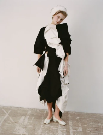 A young woman with blonde hair poses in a studio wearing a black-and-white dress, matching head covering, and white heels against a white background.
