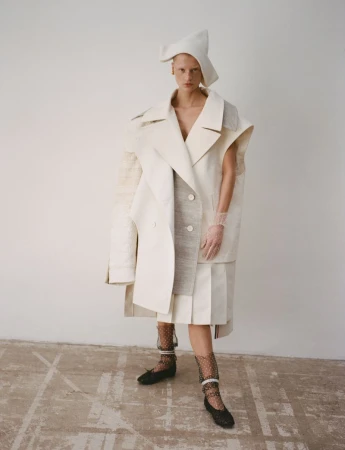 A lone fashion model in a light-toned hat and layered coat poses against a white wall on a neutral floor in a full-length fashion shot.
