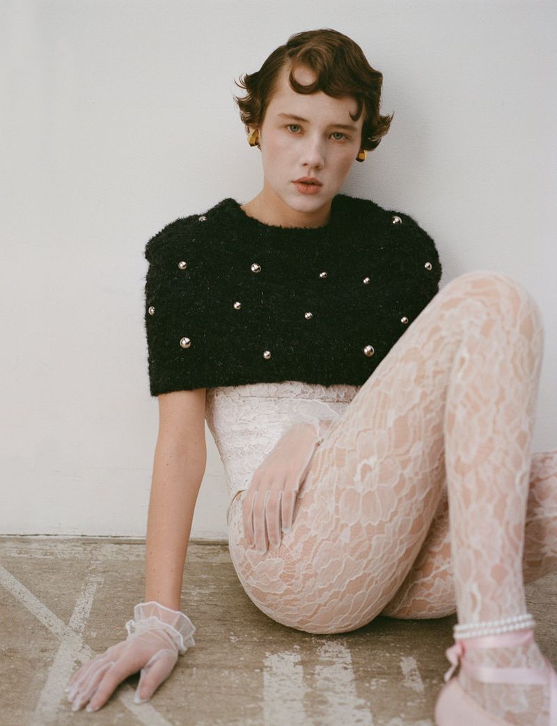 A young woman with short brown hair poses sitting on the floor in a black beaded sweater, lace leggings, and sheer gloves indoors.