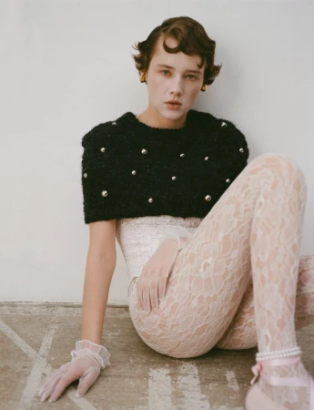 A young woman with short brown hair poses sitting on the floor in a black beaded sweater, lace leggings, and sheer gloves indoors.