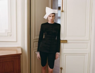 Model wearing a white sculptural hat and black suit standing in a doorway