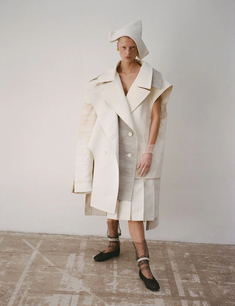 Model wearing sculptural layered white coat, pleated skirt, and unique headpiece against plain wall