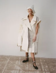 Model wearing sculptural layered white coat, pleated skirt, and unique headpiece against plain wall
