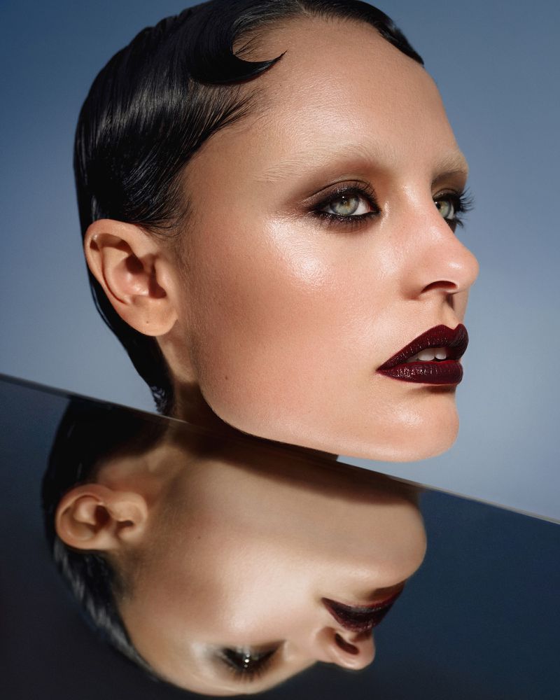 Close-up beauty shot of a woman with dark short hair, dramatic makeup, and a mirrored reflection against a gradient blue background.