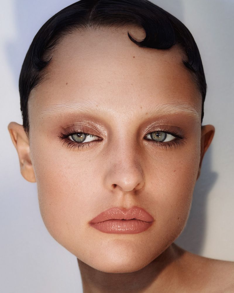 Close-up portrait of a fair-skinned woman with short dark hair, bleached eyebrows, and neutral makeup against a bright white background.