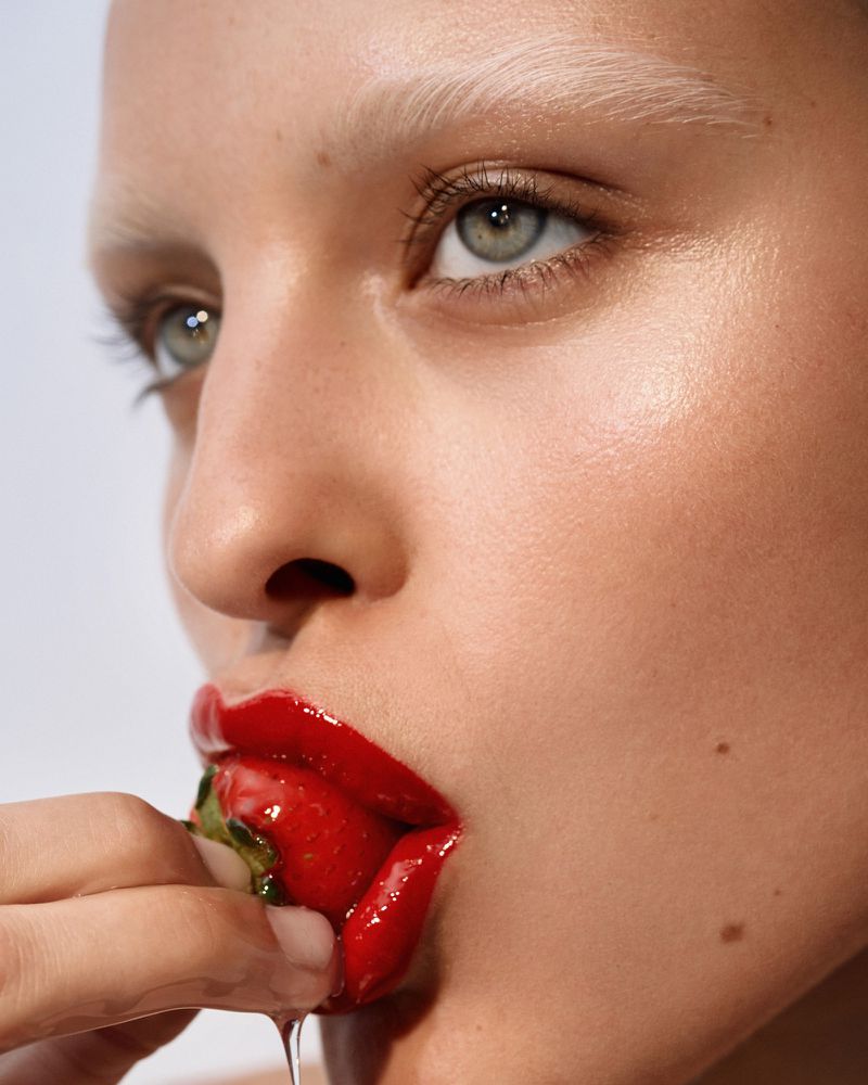 Close-up of a fair-skinned woman with bleached eyebrows holding a strawberry to her red lips against a white background.