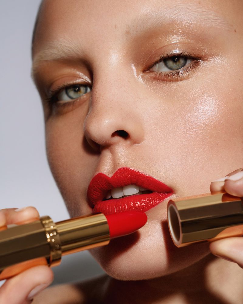 Close-up beauty shot of a young adult woman applying red lipstick in a studio, against a bright grey background.