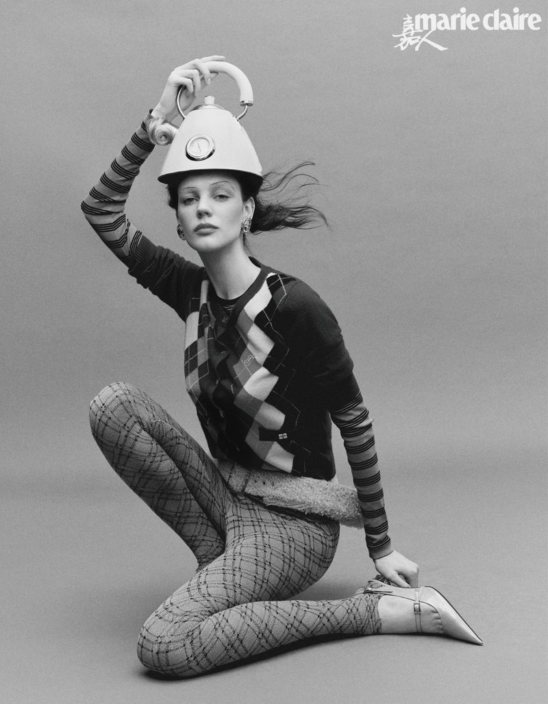 Monochrome portrait of a woman in a crouched pose wearing an argyle sweater and a kettle as a hat in a studio setting.
