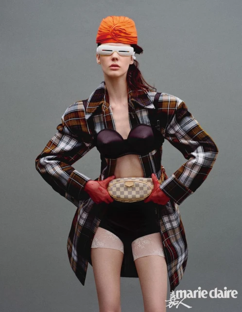 A woman poses in a brown plaid jacket, black bra and shorts, red gloves, an orange turban, and white glasses against a gray background.