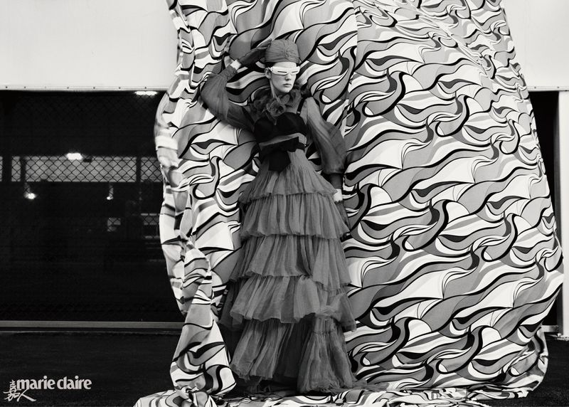 Fashion shot of a model in a tiered gown standing behind a patterned fabric, set against a dark background with visible lights.