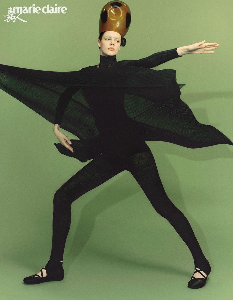 Fashion editorial image of a woman in black bodysuit, black flats, and headpiece standing against a green background. Marie Claire logo is visible.
