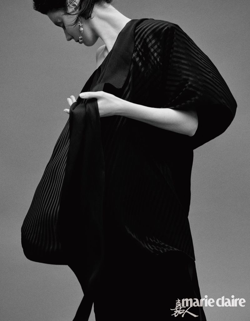 Monochrome portrait of a woman in a dark, pleated dress. She is indoors with a neutral background, accessorized with dangling earrings.