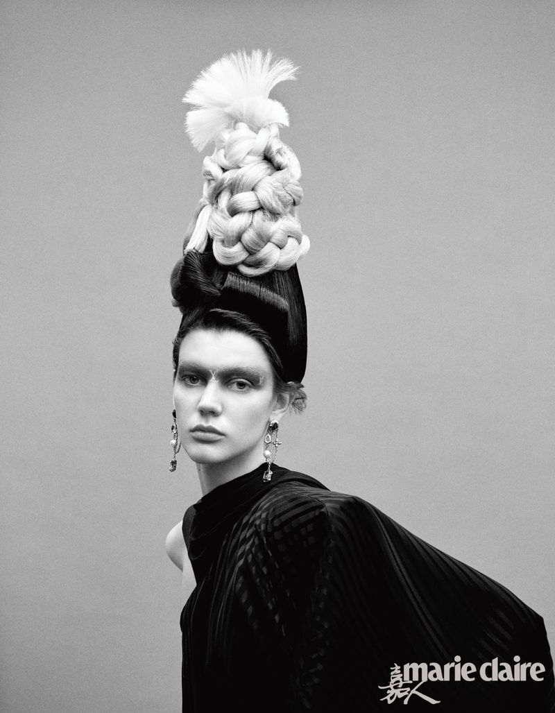 Black-and-white fashion portrait of a woman with dramatic eye makeup and an elaborate braided hairpiece, wearing a black garment against a grey backdrop.
